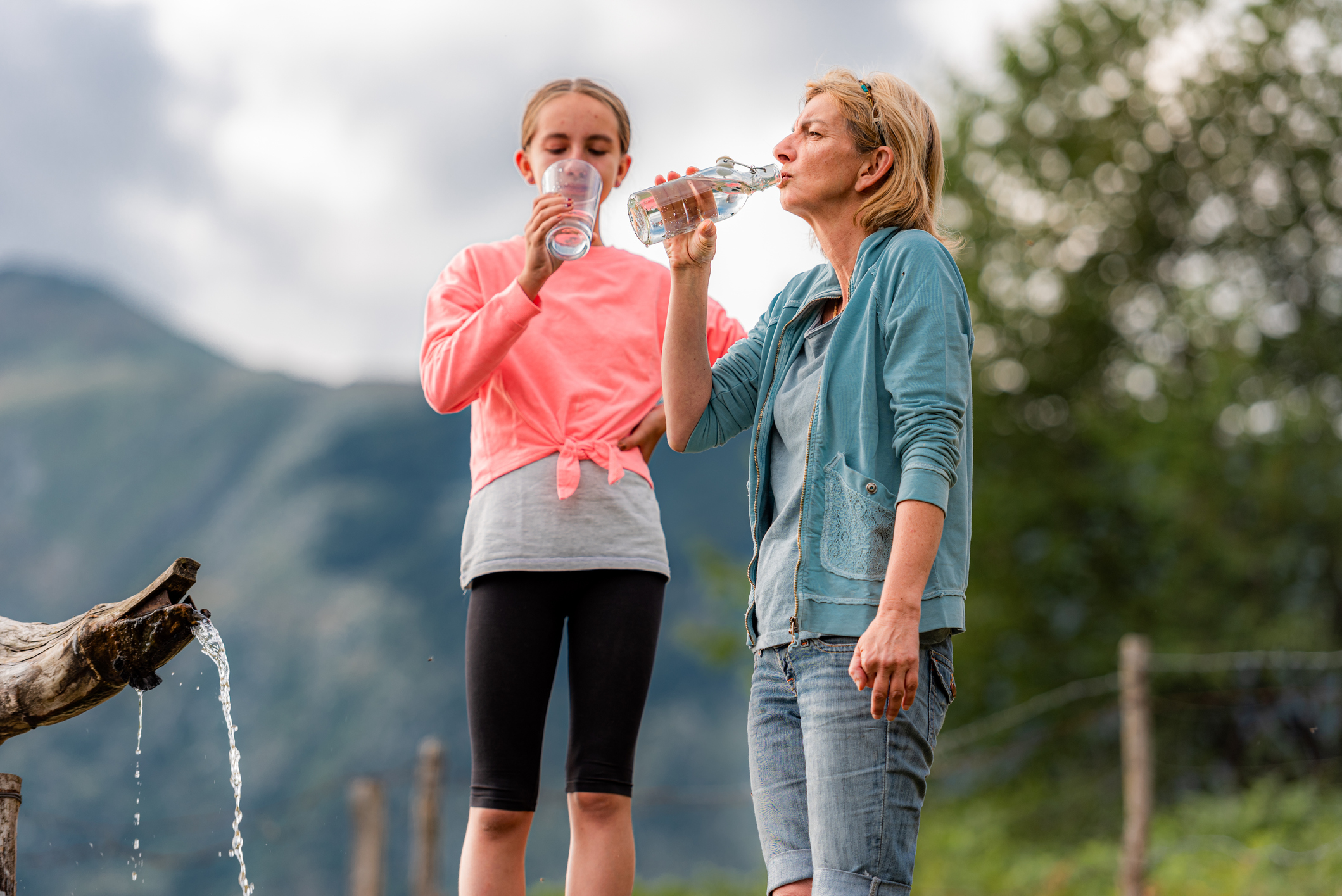 Mother and Daughter on Cloudy Summer Day Enjoy on the Pasture Admiring the View and Drinking Fresh Spring Water from Alpine Water Trough